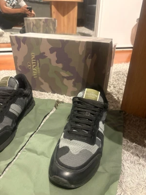 Valentino camo sneakers i svart och grått - Snygga sneakers från Valentino med camo-mönster i svart, grått och grönt. Skorna har svarta snören, mesh- och mockadetaljer samt guldlogga på plösen. Perfekta för dig som vill sticka ut med exklusiva och trendiga sneakers.