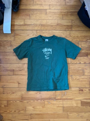 Grön Stüssy t-shirt med tryck - Säljer en grön t-shirt från Stüssy med vitt tryck på bröstet där det står flera storstäder. Klassisk passform och rund halsringning. Perfekt för dig som gillar streetwear och vill ha något stilrent men ändå med edge.