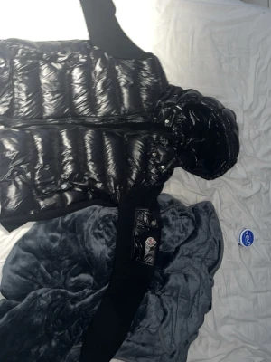 Svart dunväst med huva från Moncler - Snygg svart dunväst från Moncler med glansig finish och stickade svarta partier på sidorna. Västen har huva och en ficka med Moncler-logga på ena sidan. Perfekt för lager-på-lager och streetstyle.