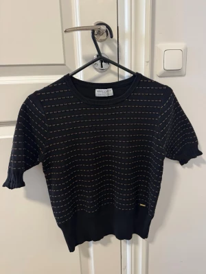 Svart stickad topp från Lindex  - Svart stickad topp  med korta ärmar och diskreta ränder i vitt och orange. Rund halsringning och ribbade muddar vid ärmslut och nederkant. 