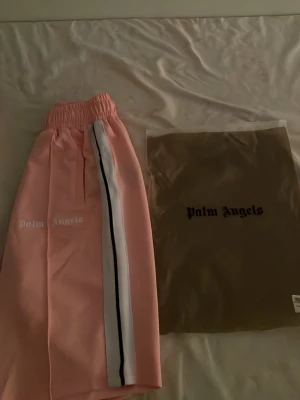 Rosa mjukisbyxor från Palm Angels - Snygga rosa mjukisbyxor från Palm Angels med vit och svart sidoremsa. Byxorna har resår i midjan och logga tryckt på benet. Perfekta för en chill och trendig look. Materialet är mjukt och bekvämt, passar perfekt till streetwear-stilen.