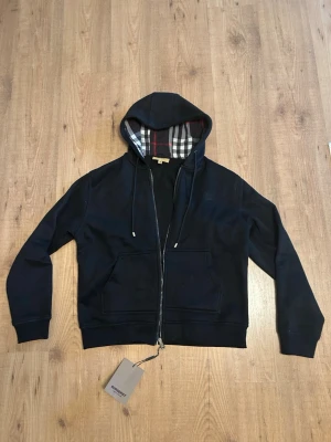 Svart hoodie från Burberry - Säljer en svart hoodie från Burberry med dragkedja och klassiskt rutigt foder i huvan. Hoodien har snören i huvan, ribbade muddar och två stora fickor framtill. Materialet är mjukt och bekvämt, perfekt för en avslappnad stil.