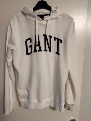 Vit hoodie från GANT med tryck - Snygg vit hoodie från GANT med stor svart logga på bröstet. Klassisk modell med huva och dragsnören, samt ribbade muddar vid ärmar och nederkant. Tillverkad i mjukt material som känns skönt mot huden. Perfekt för en avslappnad och stilren look.