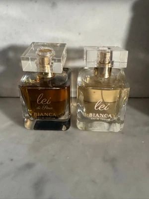 Bianca Lei Eau de Parfum duo - Två flaskor Bianca Lei Eau de Parfum, en med mörk bärnstensfärgad vätska och en med ljus gul vätska. Båda har en elegant, fyrkantig glasflaska med transparent, kubformad kork och guldiga detaljer. Perfekt för dig som gillar stilrena och lyxiga parfymer.