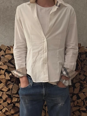 Vit långärmad skjorta från Burberry - Säljer en vit långärmad skjorta från Burberry med klassiskt rutigt mönster på insidan av manschetter och krage. Skjortan har knappar framtill och är tillverkad i bomull. Perfekt för dig som gillar stilrena plagg med diskreta detaljer.
