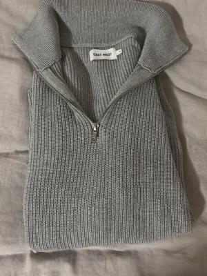 Grå stickad halvzip från East West - Snygg grå stickad tröja från East West med halvzip och hög krage. Tröjan har ribbstickad struktur och långa ärmar, perfekt för lager på lager. Enkel och stilren design som passar till det mesta.