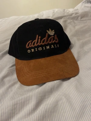 Adidas Originals svart och brun keps - Snygg Adidas Originals keps med svart tyg och brun skärm i mockaimitation. Broderad Adidas-logga och text framtill samt justerbart spänne bak. Perfekt accessoar för en sportig och avslappnad stil.