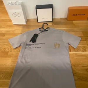 Grå t-shirt från Amiri - Säljer en snygg grå t-shirt från Amiri i 100% bomull i storlek small. Den har tryck både fram och bak med texten 'Los Angeles, California' och koordinater. Perfekt för en avslappnad stil med en touch av lyx. T-shirten är i nytt skick och har korta ärmar.