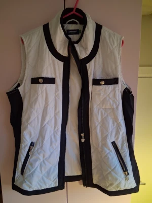 Vit och svart quiltad dunväst DAELY - Snygg quiltad dunväst från DAELY i vitt med svarta detaljer. Västen har dragkedja framtill, två fickor med dragkedja och två dekorativa bröstfickor med knapp. Perfekt för lager-på-lager och stilren look.