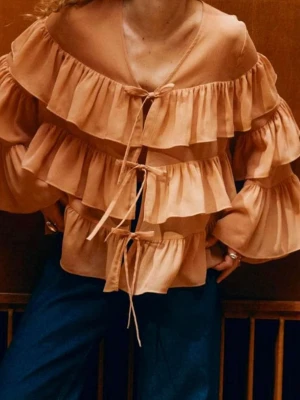 Persikofärgad volangblus  - Trendig persikofärgad blus med flera lager volanger och knytning framtill. Blusen har v-ringning och långa ärmar med voluminös passform. Tillverkad i ett lätt, transparent tyg som ger en luftig känsla.