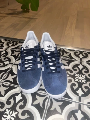 Adidas Gazelle blå sneakers - Snygga Adidas Gazelle sneakers i blå mocka med vita detaljer och klassiska tre ränder på sidorna. Låg modell med vit sula och Adidas-logga på plösen. Perfekta för en stilren och sportig look.priset kan diskuteras 