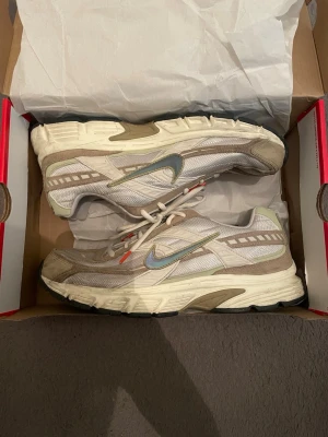 Nike Zoom Vomero 5 beige sneakers - Säljer ett par Nike Zoom Vomero 5 sneakers i beige och vitt med silverfärgad swoosh och detaljer i mesh och syntet. Skorna har en chunky sula och inslag av ljusbrunt samt små orange detaljer vid snörningen. Perfekta för dig som gillar retrostil och streetwear.