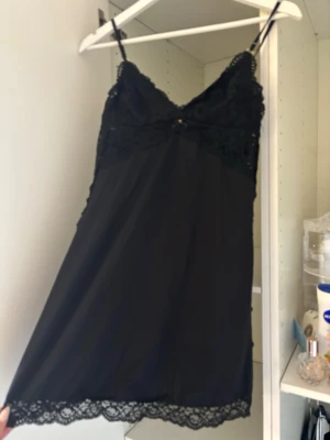 Svart spetsnattlinne med tunna axelband - Ett svart nattlinne med tunna axelband och vacker spets upptill samt längs nederkanten. Nattlinnet har en feminin V-ringning och är gjort i ett mjukt, lätt material som känns skönt mot huden. Perfekt för mysiga kvällar hemma.