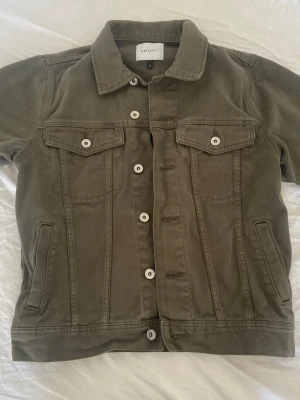 Olivgrön jeansjacka från Valiente - Säljer en olivgrön jeansjacka från Valiente med klassisk krage, vita knappar och två bröstfickor med lock. Jackan har raka linjer och snygga detaljer vid nederkanten. Perfekt för dig som vill ha en stilren och cool look.