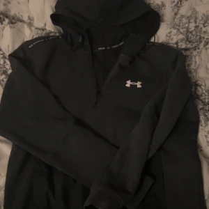 Svart hoodie från Under Armour - Säljer en svart hoodie från Under Armour med vit logga på bröstet. Hoodien har en stor huva med dragsko och ribbade muddar vid ärmslut. Perfekt för dig som gillar sportiga och bekväma kläder.