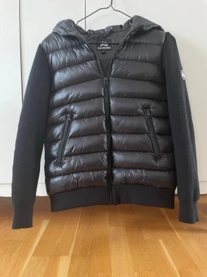 Svart cardigan jacka från Maggiore - Säljer en svart cardigan jacka från Maggiore med quiltad framsida och rygg, stickade ärmar och huva. Jackan har två dragkedjeförsedda fickor och en logotyp på ärmen. Perfekt för kalla vår och höst och riktigt snygg att bära. Kontakta vid frågor. Säljer pga kompisen köpt likadan. 