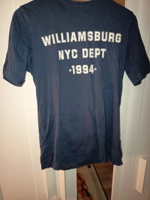 Blå Williamsburg NYC Dept t-shirt - Mörkblå t-shirt med tryck 'WILLIAMSBURG NYC DEPT 1994' och en amerikansk flagga på ärmen. Skön och avslappnad passform, perfekt till jeans eller shorts. Klassisk college-stil med korta ärmar.