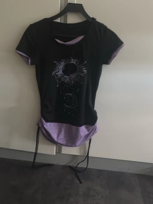 Svart och lila topp med soltryck - Cool svart topp med lila detaljer och tryck av sol och måne framtill. Toppen har korta ärmar, rund hals och snörning i sidorna för en extra touch. Perfekt för dig som gillar unika detaljer och kontrastfärger.