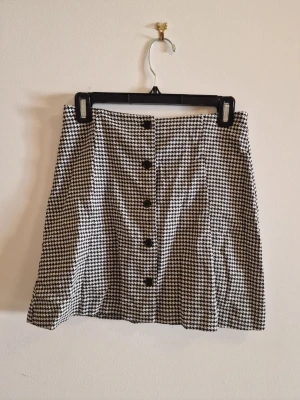 Houndstooth kjol från Monki - Snygg svartvit kjol från Monki i klassiskt hundtandsmönster. Kjolen har knappar framtill och en lätt A-linjeformad siluett. Perfekt för dig som gillar retrovibbar och vill sticka ut med ett trendigt mönster.