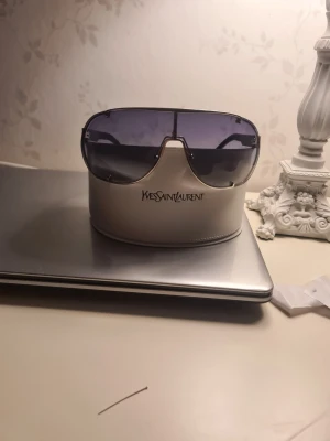 Svarta solglasögon från Yves Saint Laurent - Säljer ett par snygga svarta solglasögon från Yves Saint Laurent. De har en klassisk rund form och kommer med ett vitt, exklusivt fodral med logga. Perfekta för dig som vill ha en tidlös accessoar med lyxig känsla. glasögonen har några repor på linserna men ser bra ut