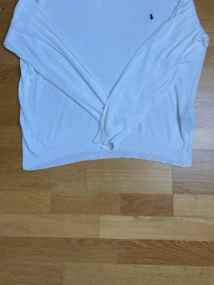 Vit stickad tröja från Polo Ralph Lauren - Vit stickad tröja från Polo Ralph Lauren med V-ringning och diskret broderad logga i mörkblått på bröstet. Tröjan har långa ärmar och ribbstickade muddar vid ärmslut och nederkant. Perfekt för en clean och klassisk stil.