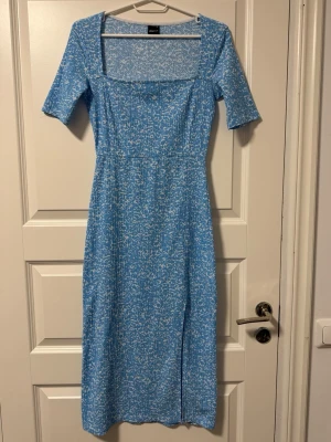 Blå långklänning med mönster - Ljusblå långklänning med vitt småmönster och fyrkantig ringning. Klänningen har korta ärmar och en smickrande passform.