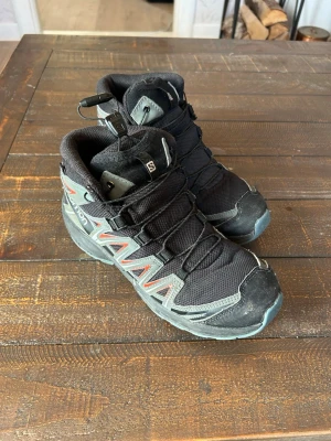 Svarta kängor från Salomon, 38 - Säljer ett par svarta Salomon XA PRO kängor med grå och röda detaljer. Skorna har hög modell, robust sula och snabbsnörning, strl 38. Obs! Ena snöret är på väg att gå av, se bild