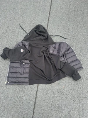 Svart Moncler väst med huva - Säljer en svart Moncler väst med huva och dragkedja. Västen har vadderade paneler på sidorna och en mjuk sweatshirtliknande baksida. Klassisk Moncler-logga på ärmen och dragsko i huvan. Perfekt för lager-på-lager och streetwear. Är xxl i barn storlek passar 14årikg 