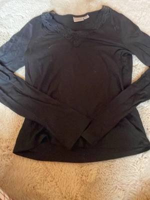 Svart långärmad topp med spets - Svart långärmad topp från Gina Tricot med snygg spetsdetalj runt v-ringad hals. Materialet är mjukt och stretchigt, perfekt för att matcha med jeans eller kjol. Enkel och stilren design som passar till många olika outfits.