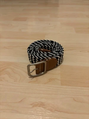 Svartvitt randigt bälte med läderdetalj - Snyggt svartvitt bälte med randigt mönster och silverspänne. Bältet har bruna läderdetaljer vid spännet som ger en stilren kontrast. Perfekt accessoar för att lyfta din outfit.