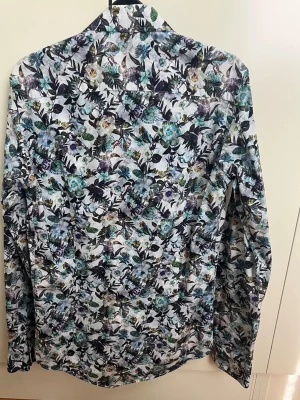 Blommig skjorta från Sand - Snygg skjorta från Sand med blommigt mönster i blått, grönt och lila på vit botten. Skjortan har lång ärm, klassisk krage och knäppning framtill. Perfekt för dig som vill sticka ut med en färgglad och trendig look.
