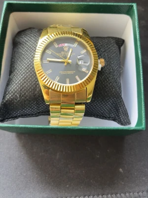 Rolex guldfärgad klocka Day-Date - Stilren guldfärgad Rolex Day-Date med svart urtavla och dag- samt datumvisning. Klockan har en klassisk länk i metall och markerade index. Perfekt accessoar för dig som vill sticka ut med en lyxig look.