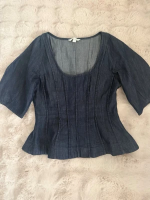 Mörkblå denim blus med vida ärmar - Snygg mörkblå blus i denim-look med markerad midja och vida, halvlånga ärmar. Toppen har en djup rundad ringning och sömmar som ger en figurnära siluett. Perfekt för dig som vill ha en trendig och avslappnad stil.