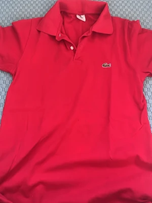 Röd pikétröja från Lacoste - Klassisk röd pikétröja från Lacoste med den ikoniska lloggan broderad på bröstet. Tröjan har korta ärmar, krage och knappar vid halsen. Perfekt för en stilren och somrig look.