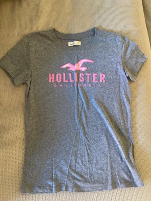 Grå Hollister t-shirt med rosa tryck - Snygg grå t-shirt från Hollister med klassiskt rosa logotyp-tryck och texten 'California' framtill. T-shirten har rund halsringning och korta ärmar. Tröjan har använts en del.