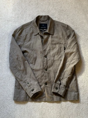 Beige/Grön overshirt från Junk de Luxe - Stilren beige overshirt från Junk de Luxe i linnemix. Klassisk krage, knappstängning framtill och en bröstficka. Perfekt lager-på-lager-plagg med avslappnad passform och snygga detaljer vid ärmslut.