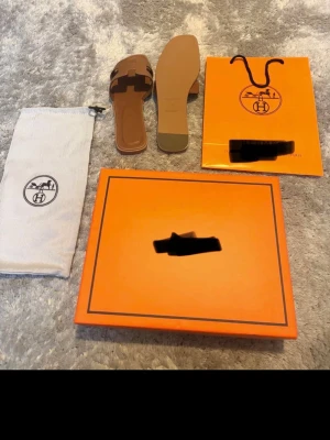 Hermè Bruna Oran sandaler  - Säljer ett par bruna Oran sandaler i läder med den ikoniska H-remmen. Sandalerna har öppen tå, platt sula och fyrkantig form. Kommer med original ask, dustbag och påse. Perfekta för en trendig och stilren look.