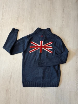 Mörkblå stickad tröja från Bondelid - Mörkblå stickad tröja från Bondelid med hög krage och knappar. På ryggen finns en stor Union Jack-flagga i rött, vitt och blått. Framtill broderad logga och text. Tröjan har långa ärmar och ribbade muddar. Perfekt för dig som gillar brittisk stil.