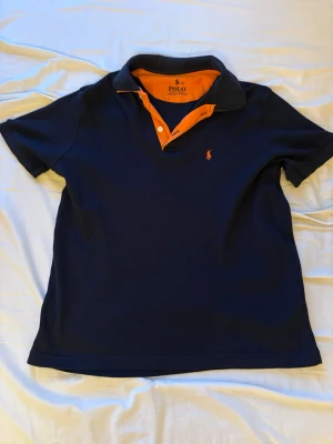 Mörkblå pikétröja från Polo Ralph Lauren - Snygg mörkblå pikétröja från Polo Ralph Lauren med orange detaljer på insidan av kragen och knappraden. Klassisk polokrage och korta ärmar. Ikoniska loggan broderad i orange på bröstet. Liten i storleken så den är inte som en XL