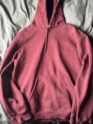 Mörkröd hoodie med magficka - En stilren mörkröd hoodie med huva och dragsko. Hoodien har en klassisk känguruficka framtill och ribbade muddar vid ärmslut och nederkant. Perfekt för en avslappnad look och enkel att matcha med jeans eller joggers.