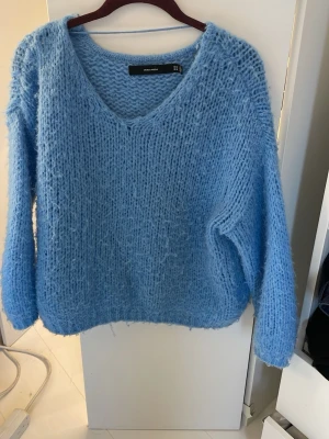 Ljusblå stickad tröja Vero Moda - Mysig ljusblå stickad tröja från Vero Moda med v-ringning och lång ärm. Tröjan har en fluffig, lite lösstickad struktur som ger en avslappnad look. Perfekt för att hålla värmen under kyliga dagar.