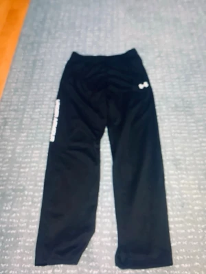 Svarta mjukisbyxor från Under Armour - Säljer ett par svarta mjukisbyxor från Under Armour med vitt logotryck på låret och texten UNDER ARMOUR längs benet. Byxorna har rak passform och resår i midjan. Perfekta för chill eller träning.