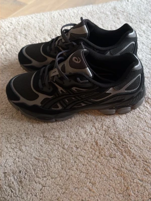 Asics Graphite GEL NYC - Snygga sneakers från Asics i svart och grått med meshpaneler och detaljer i syntetmaterial. Skorna har en sportig design med lågt skaft och klassisk snörning. Perfekta för dig som gillar en stilren och bekväm look.