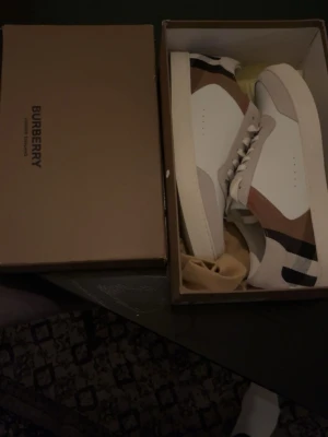 Burberry vita sneakers med rutigt mönster intresse koll ge bud! - Snygga vita sneakers från Burberry med klassiskt beige, svart och rött rutmönster på sidan. Skorna har låga kanter, vita snören och en stilren design i läder och textil. Perfekta för dig som vill ha en exklusiv touch på din outfit. Ge bud osv har användt dom efter ja tog bilden men de är nästan som nya.