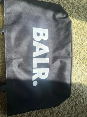 Svart väska från BALR. - Säljer en svart väska från BALR. med stor vit logga på framsidan. Väskan är tillverkad i slitstarkt tygmaterial och har en enkel, rektangulär form. Perfekt för dig som gillar stilrena accessoarer med sportig känsla. Jag had aldrig använt den