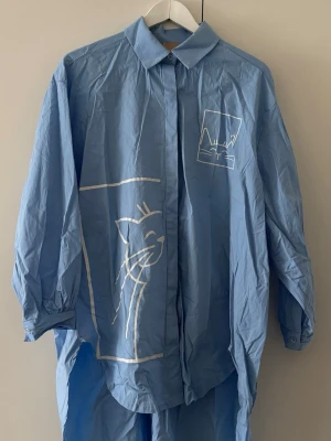 Blå skjortklänning med kattmotiv - Oversized blå skjortklänning med långa ärmar och vit kattillustration framtill. Klänningen har klassisk krage, knäppning hela vägen och är tillverkad i ett lätt bomullstyg. Perfekt för dig som gillar unika och lekfulla detaljer.