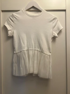 Vit topp med volangdetalj - Snygg vit topp med korta ärmar och rund halsringning. Nedre delen har en volang i tunt skick. Perfekt att styla med jeans eller kjol.