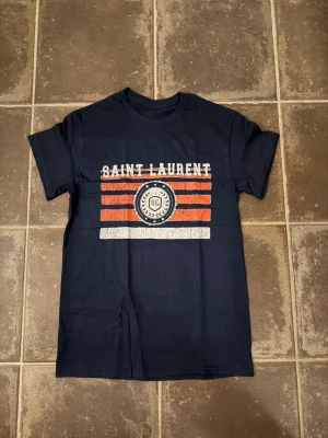 Saint Laurent t shirt  - Riktigt fet och eftertraktad t-shirt med Saint Laurent loggan i storlek S. Väldigt bra skick!  Obs: Jag är lite osäker på äktheten då lappen i nacken saknas, därför säljs den billigare (499 kr, nypris ca 3000 kr).  längd cirka 65cm, bredd cirka 44cm. Tveka inte att höra av dig vid frågor eller funderingar!