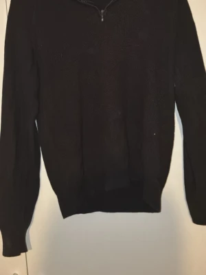Svart stickad tröja med halv dragkedja från Dressmann - En enkel svart stickad tröja med halv dragkedja vid halsen. Tröjan har långa ärmar och ribbade muddar vid ärmslut och nederkant. Perfekt för lager på lager och en clean look.