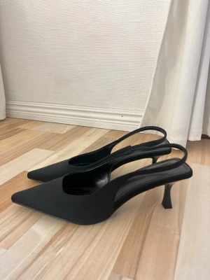 Svarta spetsiga slingback-pumps - Stilrena svarta pumps med spetsig tå och smal klack. Skorna har öppen häl med slingback-rem och är tillverkade i ett matt material. Perfekta för dig som gillar en klassisk och trendig look. Oanvända endast testade!!!!!